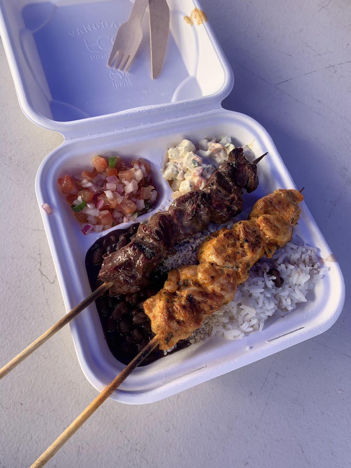 A prato feito box with beef and chicken skewers, black beans, rice, salsa and potato salad